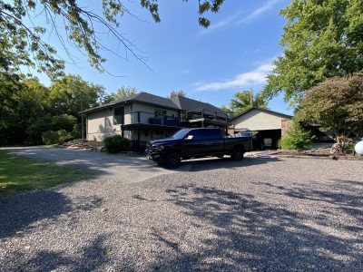 House with a black pickup parked on a gravel driveway, surrounded by trees under a blue sky.