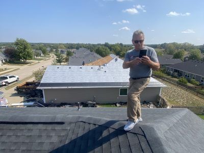 Person standing on a shingled roof, looking at a phone, with a suburban neighborhood in the background.
