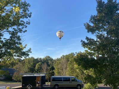 Hot air balloon over trees and parked van under a blue sky