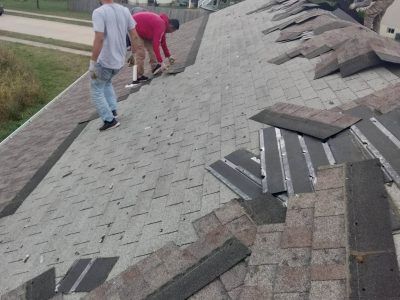 Workers installing roof shingles on a sloped roof, with loose materials laid out nearby.