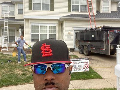 Man in red cap and blue sunglasses taking selfie with house, worker, and dump truck in background