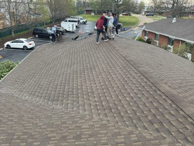 People on a shingled roof beside parked vehicles in a residential driveway