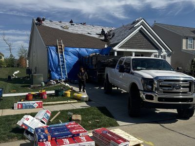 Workers on a house roof with a blue tarp and ladder, tools scattered on the driveway near a white truck.