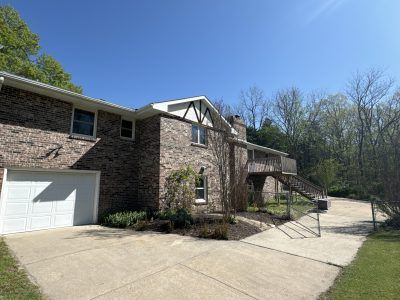 Stone house with white garage door and driveway under a clear blue sky, beside wooded hills