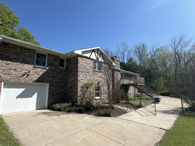 Brick house with a white garage, driveway, and wooded hillside under a clear blue sky