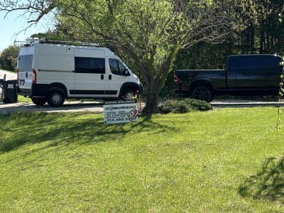White van and black pickup parked on grass beside a tree with a small yard sign