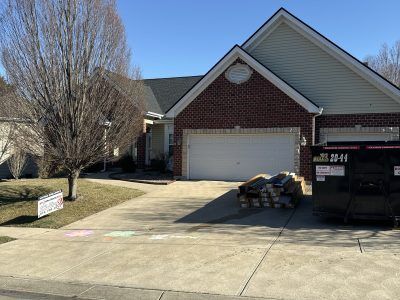 Suburban brick house with white garage, driveway, leafless trees, and a dumpster in front