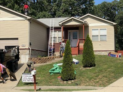 Workers on a house roof and porch during exterior repairs, with ladders, tools, and a dumpster in the yard