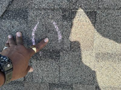 Hand pointing at a purple chalk mark on rough gray pavement beside a large painted arrow shadow