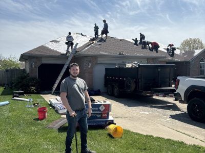 People on a roof repairing storm damage beside a driveway and garage, with tools and debris below.