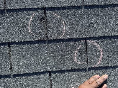 Close-up of asphalt roof shingles with chalk-marked circles and a person’s hand at the bottom right