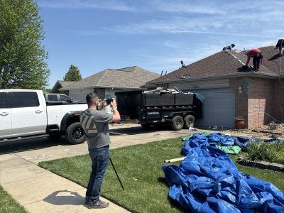 Person filming a roof repair beside a house, with a truck, trailer, and blue tarp-covered debris in the yard
