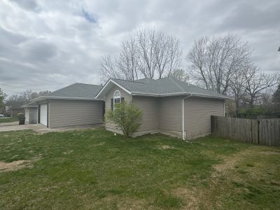 Single-story beige house with attached garage, gray roof, and a fenced backyard under an overcast sky