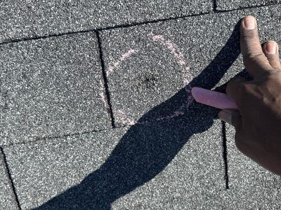 Hand tracing a chalk outline on gray asphalt or shingle surface, casting a long shadow.