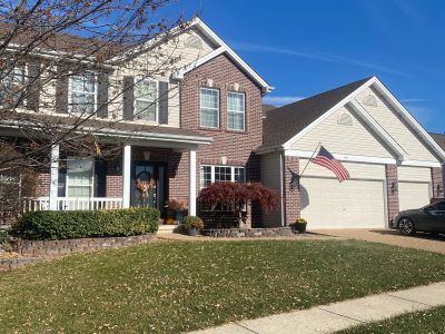Suburban brick-and-beige house with a front porch, garage, lawn, and American flag on a clear day