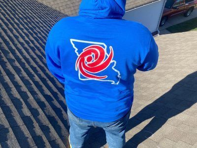 Person in a blue hoodie with a red-and-white spiral logo, standing outdoors on a sunny paved surface.
