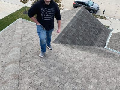 Person standing on a shingled roof beside a dormer, viewed from above.