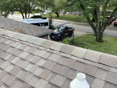 Roof shingles with a white vent cap; below, a black truck and white van parked beside a tree-lined street.