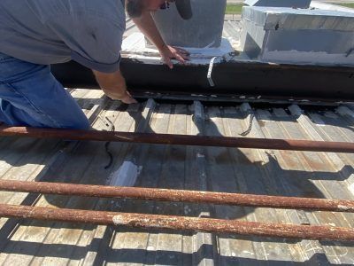 Worker kneels on a rooftop, applying waterproof sealant around a vent beside weathered metal pipes.