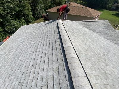 Worker on a shingled roof inspecting the ridge near a house in a suburban neighborhood