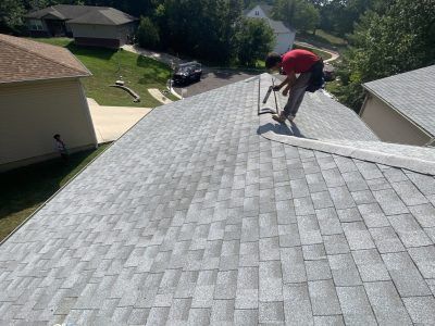 Person kneeling on a gray shingled roof with a tool, inspecting repairs in a suburban neighborhood