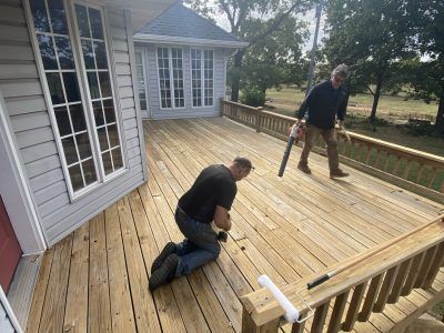 Two people working on a wooden deck beside a house, one kneeling and one standing with tools.