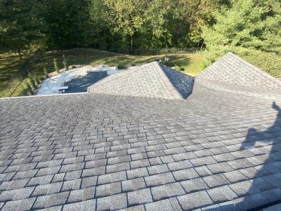 Aerial view of a gray shingled roof with two peaks beside trees and a fenced yard