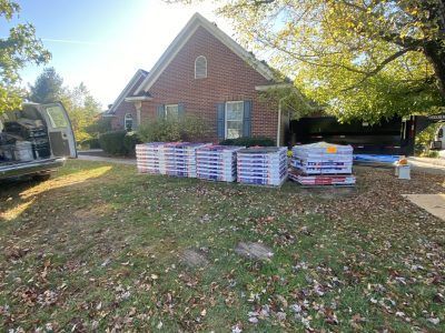 Brick house with stacked lumber and building materials in the front yard, surrounded by trees and a parked vehicle