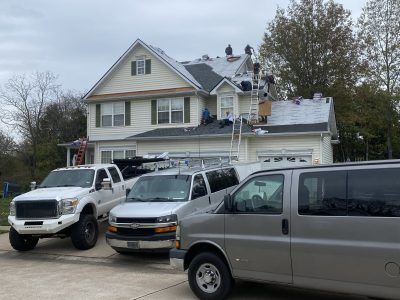 Parked vehicles in front of a large beige house with a sloped roof on a cloudy day
