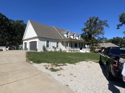 White house with a steep roof and driveway, seen on a sunny day with parked vehicles and trees.