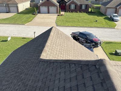 Aerial view of a suburban house roof, driveway, and parked black car on a quiet street.