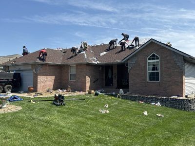 Workers repairing a brick house roof with debris scattered across the lawn and a clear blue sky.