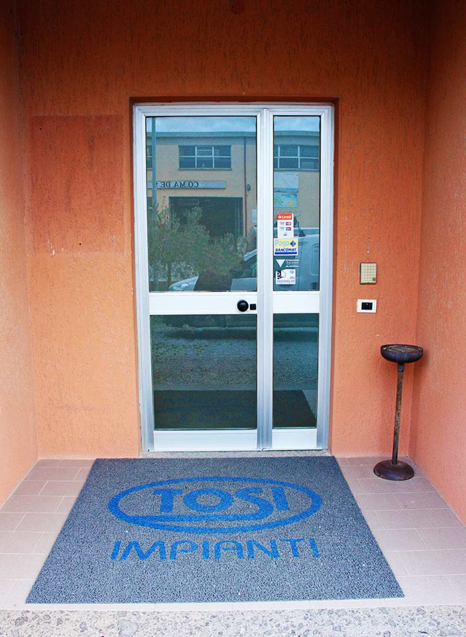 Ingresso di Tosi Impianti con zerbino grigio con logo del marchio davanti a una porta a vetri incastonata in una parete color pesca.