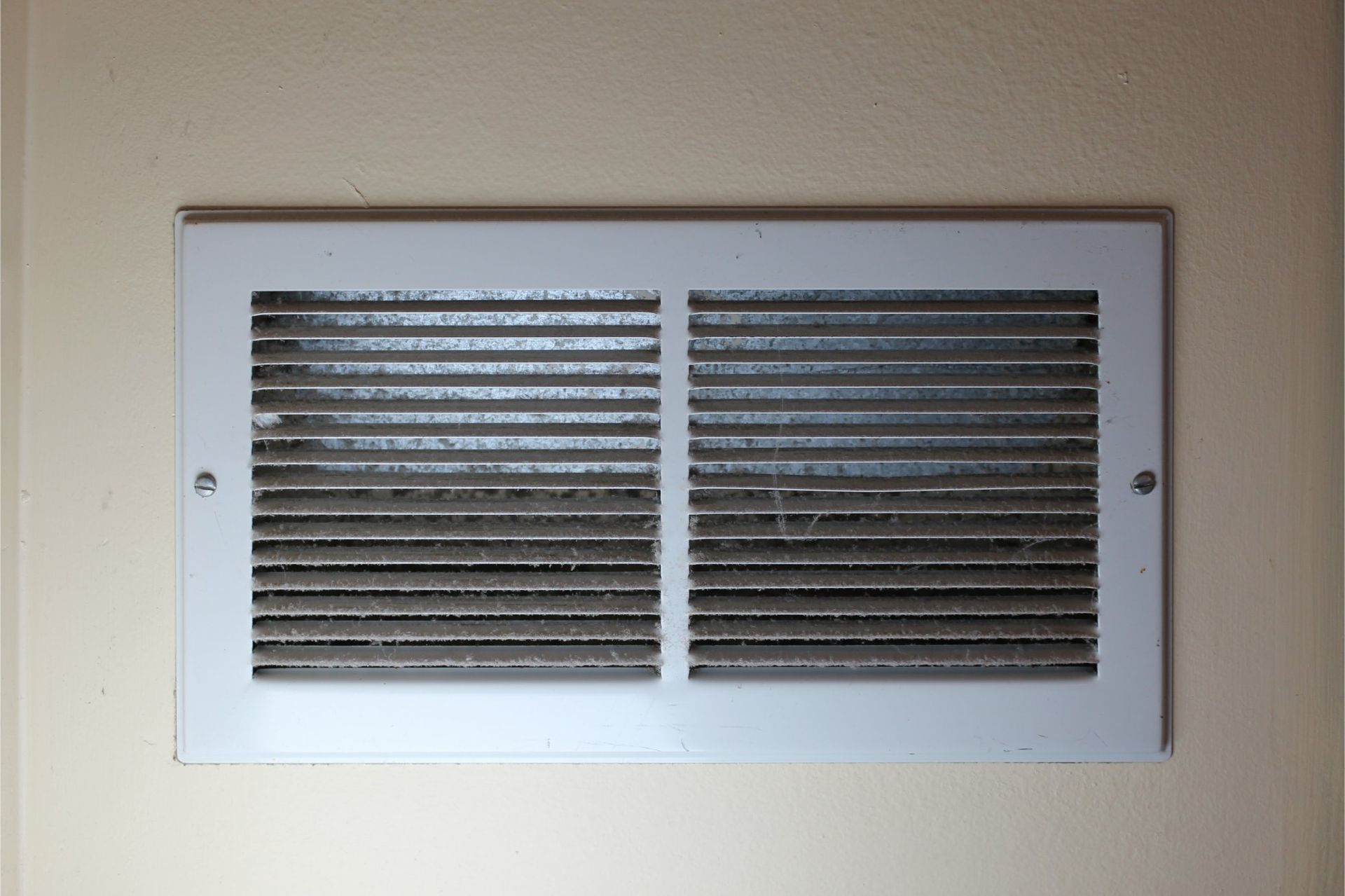 White, rectangular air vent in a beige wall, filled with dust and debris.