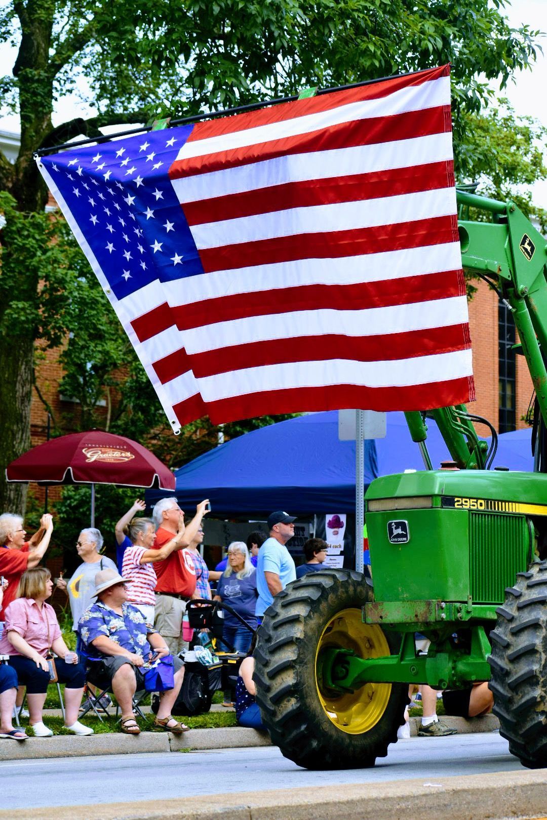 Large American flag on a green tractor at a parade with seated spectators.