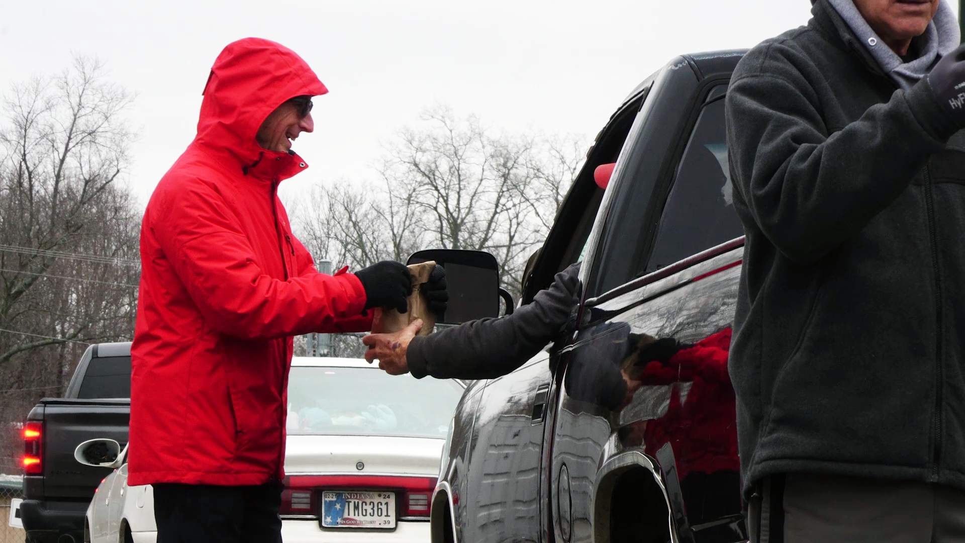 Person in a red hooded jacket shakes hands with someone beside a black truck in a snowy parking lot.