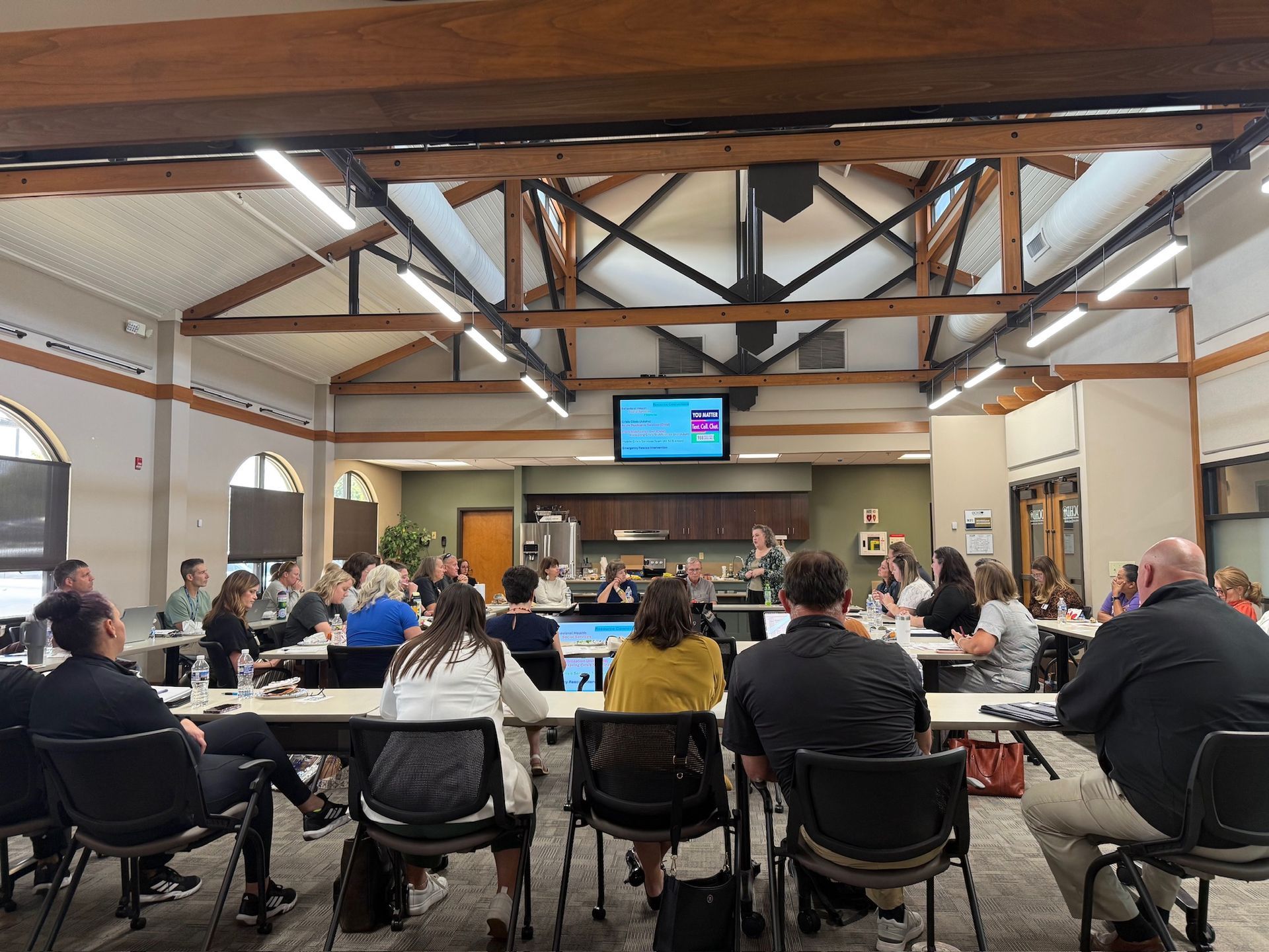 People seated at tables in a hall, watching a presentation on a wall-mounted screen.