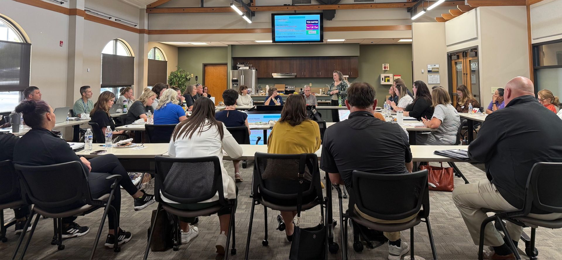 Audience seated at tables in a meeting room, facing the front under bright ceiling lights.