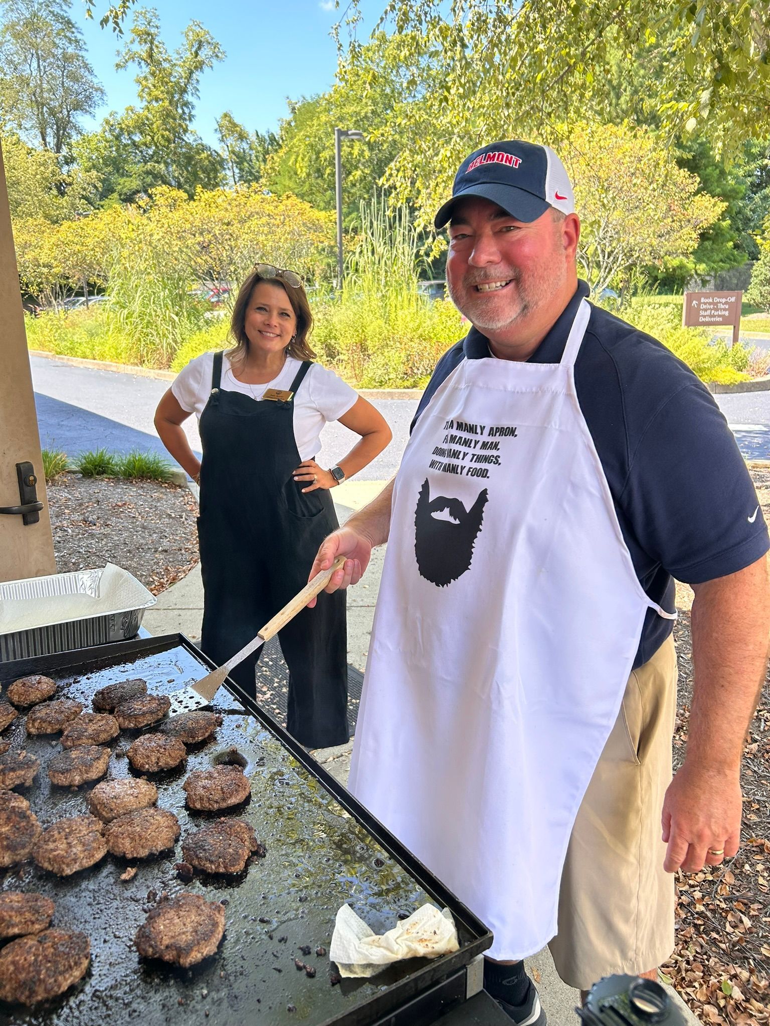 Two people grilling burgers at an outdoor barbecue, with one smiling by the grill and trees in the background