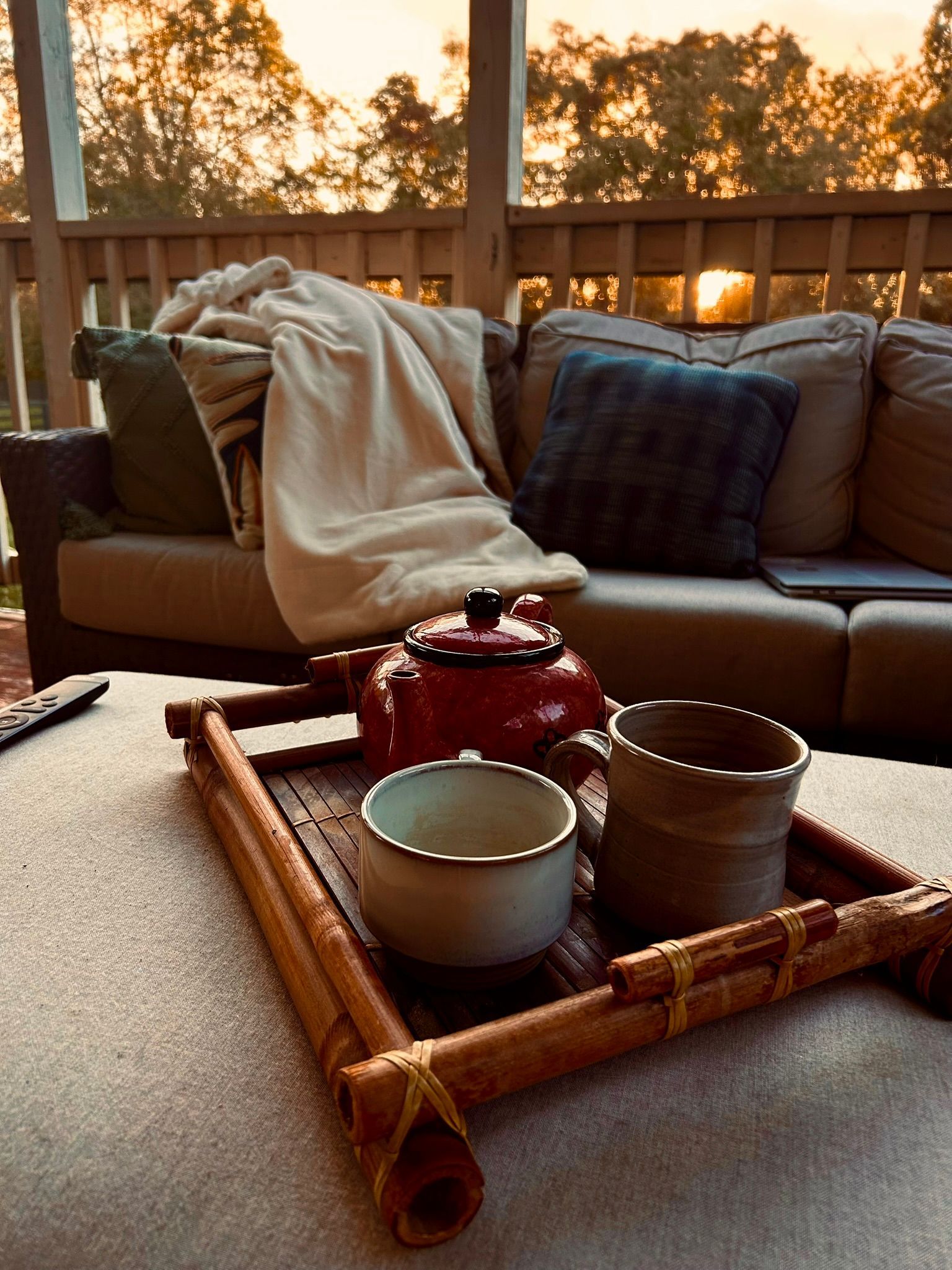 Cozy patio scene at sunset with a tray of teapot and cups on a table beside a couch and blanket.