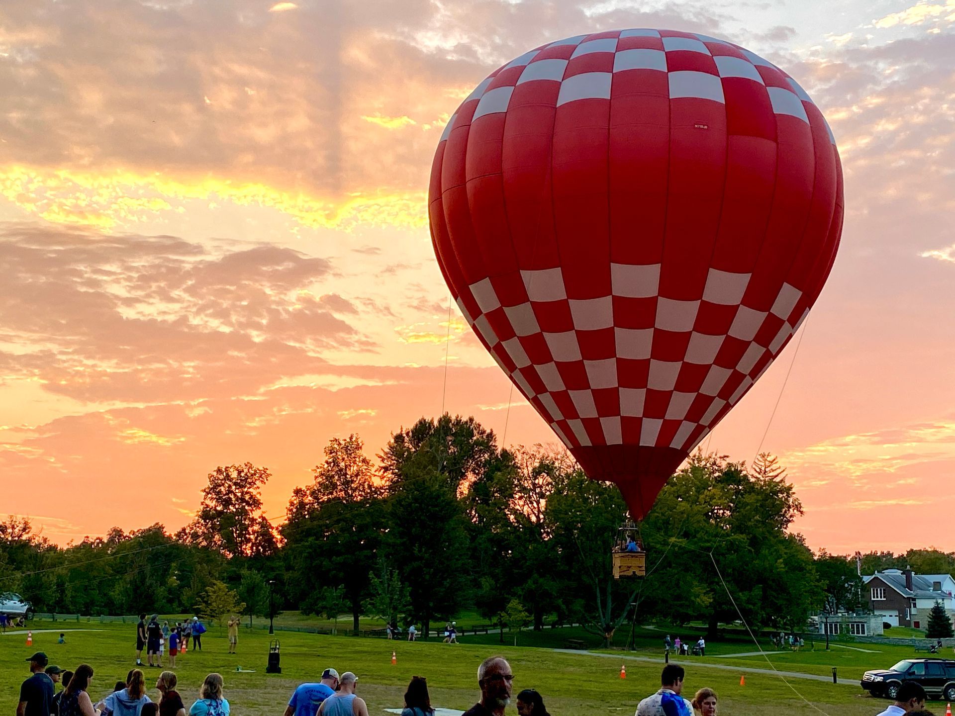 Red checkerboard hot air balloon over a grassy field at sunset with people watching nearby