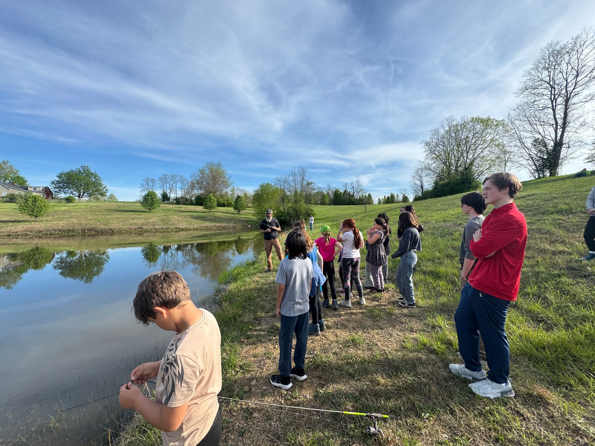 People standing by a pond in a grassy park, looking into the water under a bright blue sky.