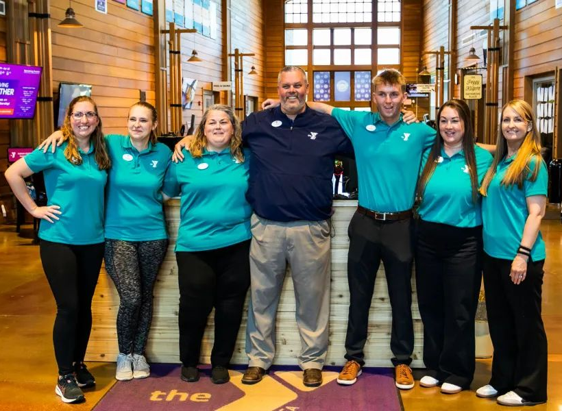 Group of six people in teal shirts posing indoors with a man in a navy shirt at center.