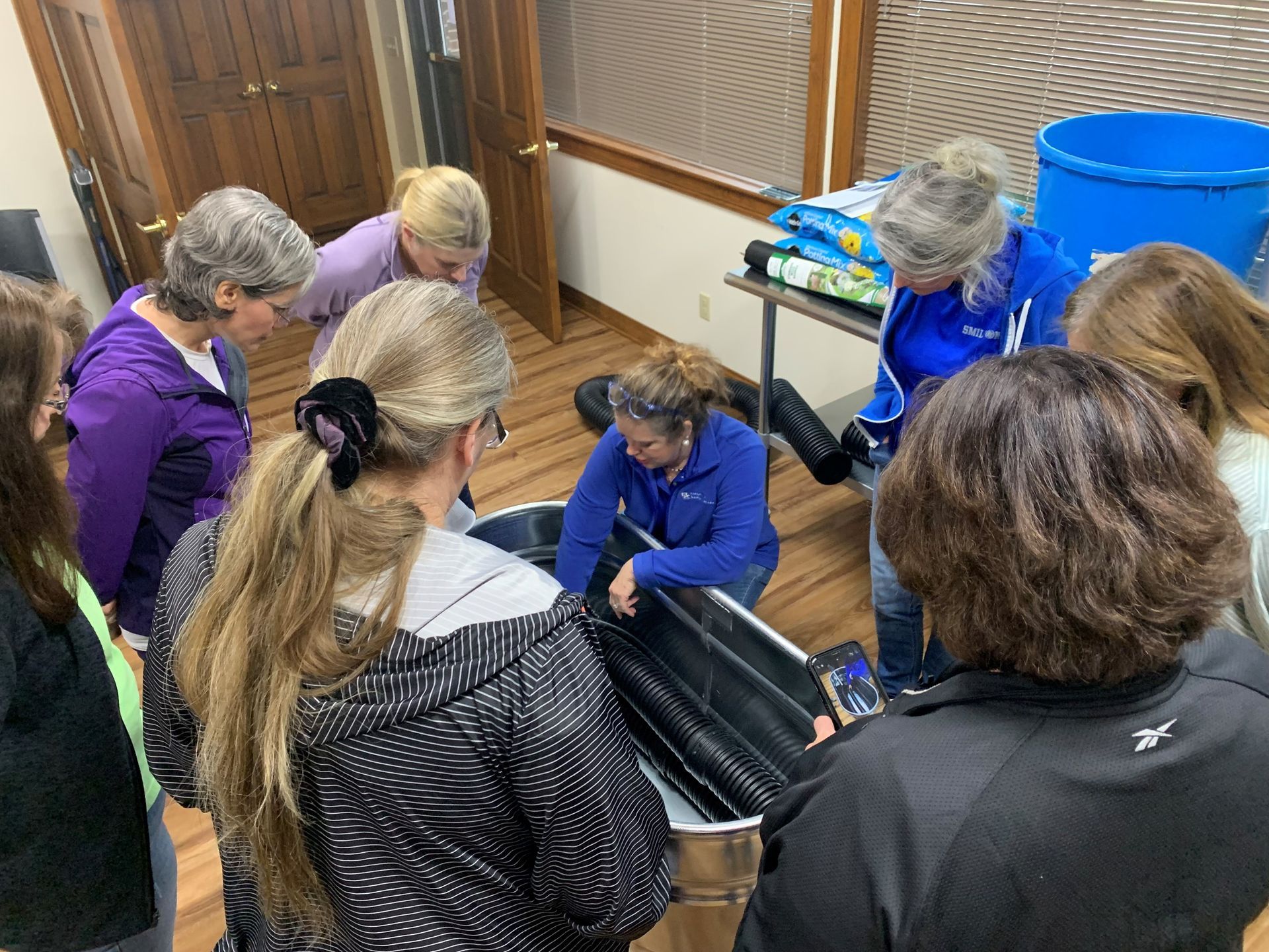 Group of people in hairnets and gloves sorting items around a table in a room with blue bins and blinds.