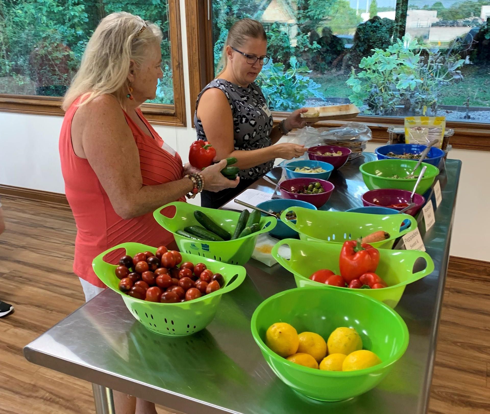Two people sample produce at a colorful market table with bowls of tomatoes, lemons, and peppers.