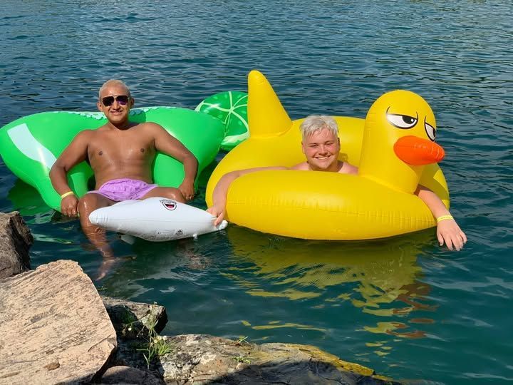 Two people float on colorful inflatable swans in clear blue water near a rocky shore.