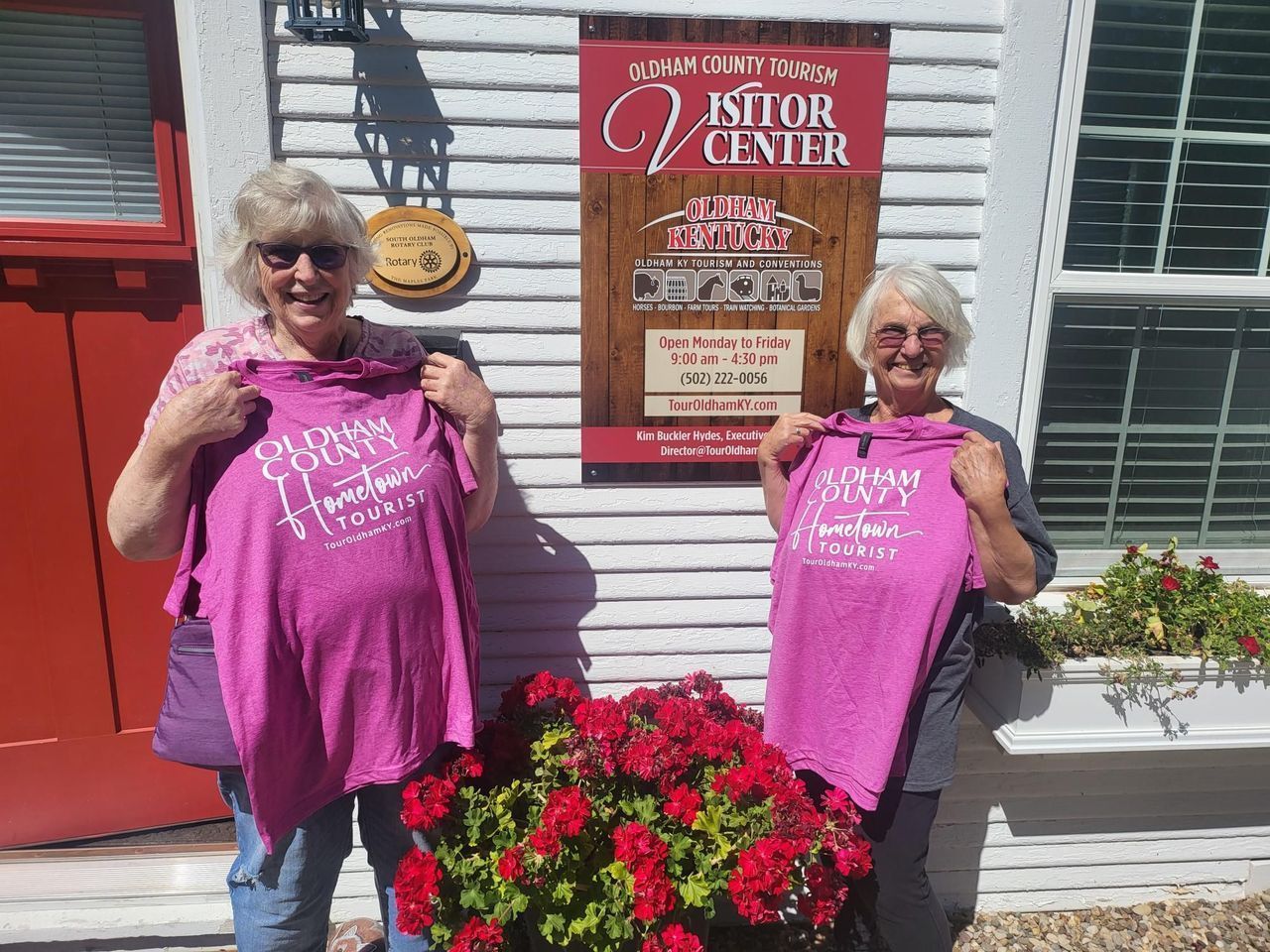 Two women holding matching pink shirts in front of a flower planter and a red storefront sign