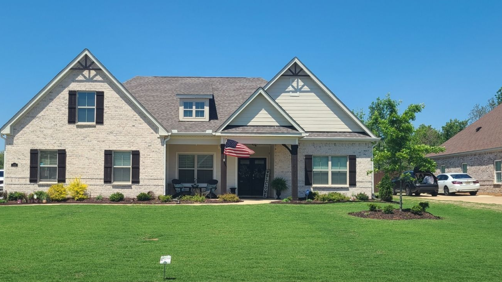 A one-story light-colored brick house with a gabled roof, shutters, and a front porch under a clear blue sky.