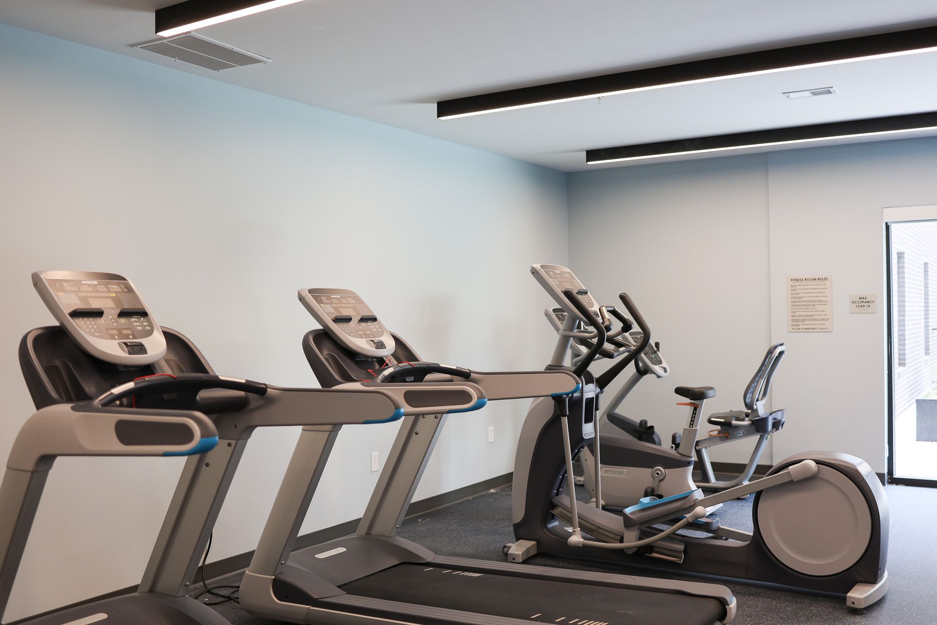 3-Bedroom Affordable Apartments in Downtown Lincoln, NE - Union at Antelope Valley - Fitness Center With Treadmills, Elliptical, Stationary Bike, Modern Linear Lights, And Glass Entry Door