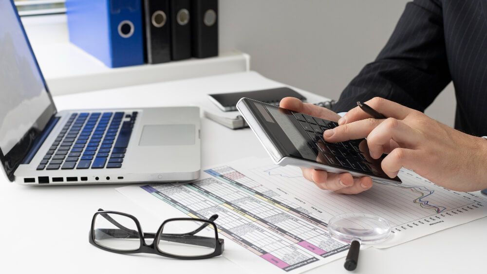 A person is using a calculator in front of a laptop computer.